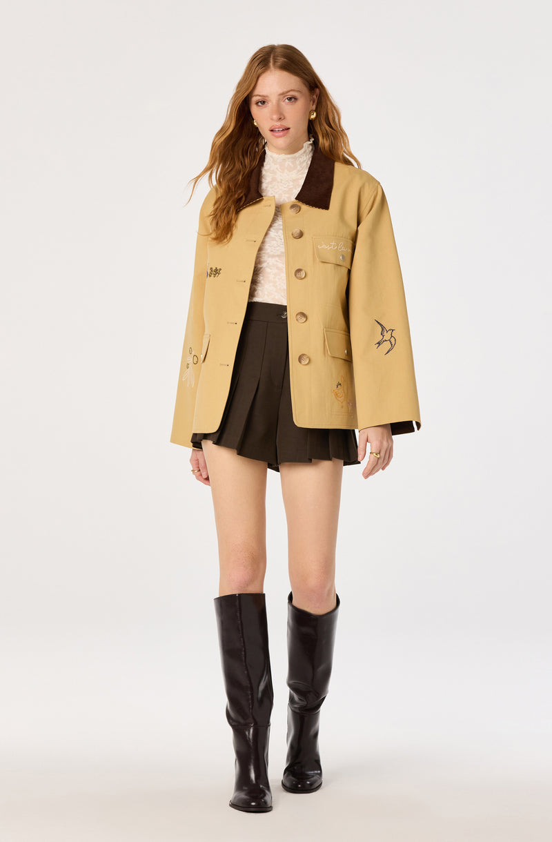 Person wearing a beige jacket with embroidered designs, black pleated shorts, a light lace top, and tall black boots.