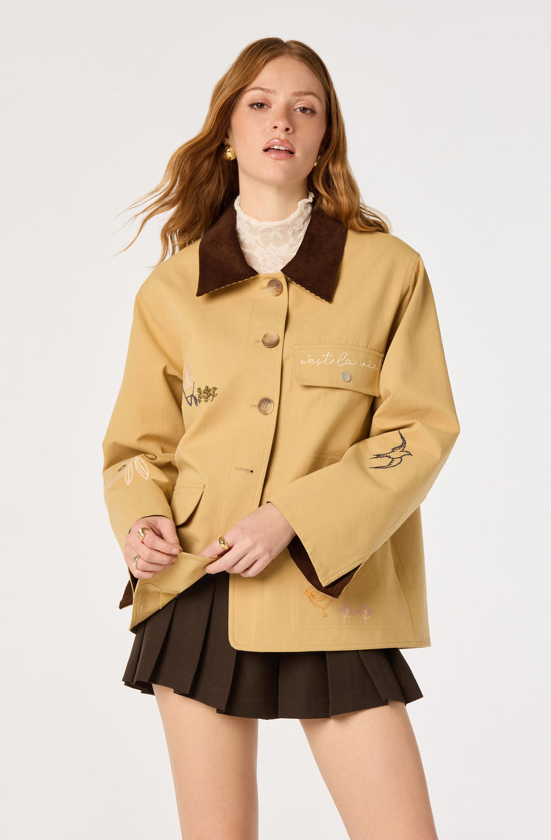Woman wearing a beige jacket with embroidered details and a dark brown collar, paired with a dark brown pleated skirt.