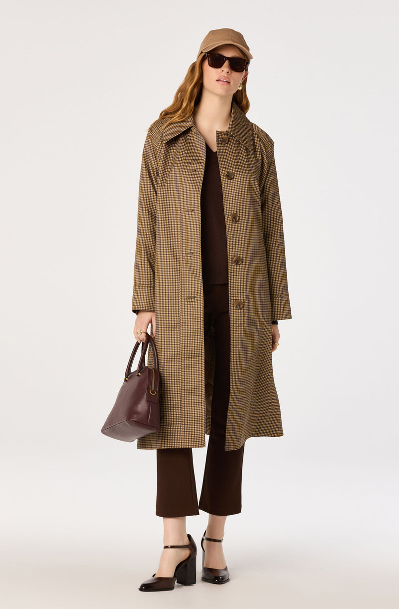 Woman in brown checked trench coat, brown pants, black heels, tan cap, and sunglasses, holding a dark brown handbag.