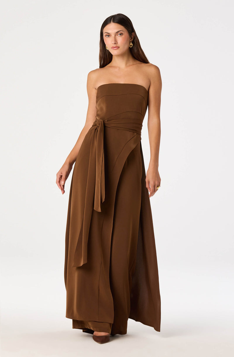 Brown strapless jumpsuit with a large fabric bow on the waist and wide-leg pants.