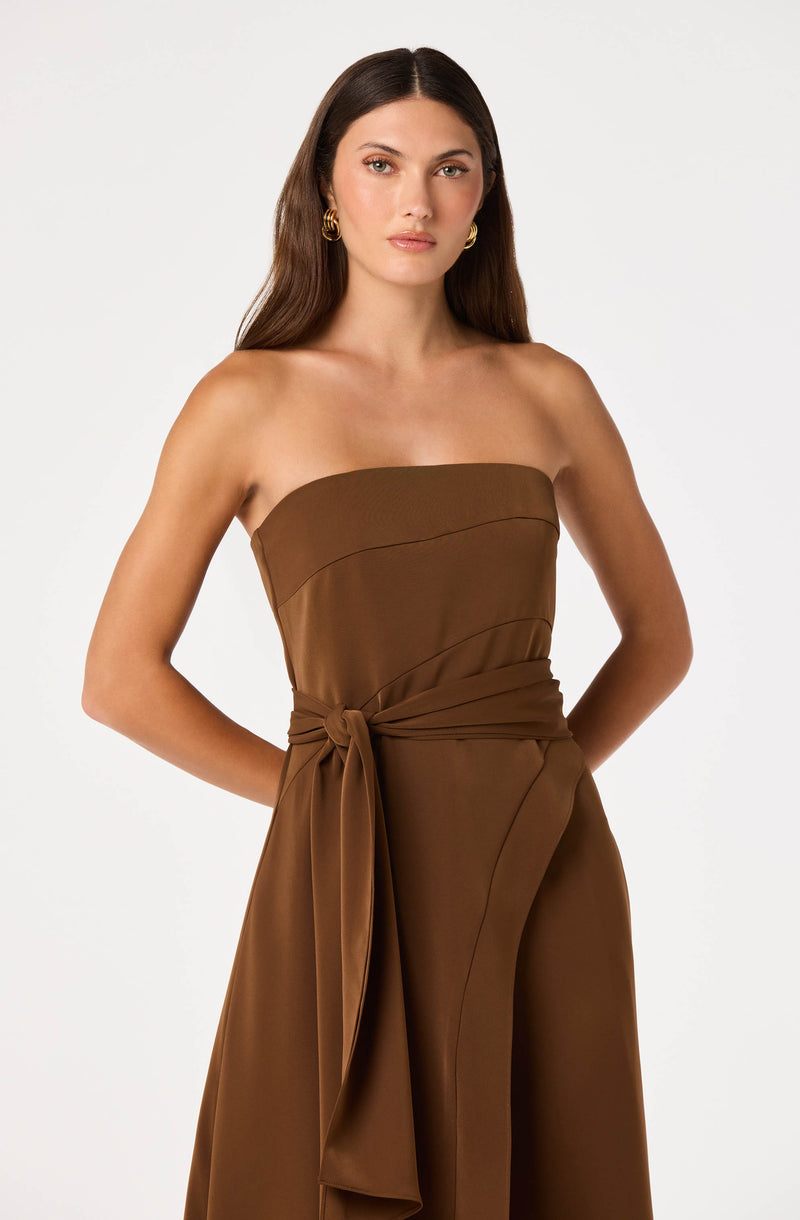 Woman in a strapless brown jumpsuit with a tied waist, standing against a white background.