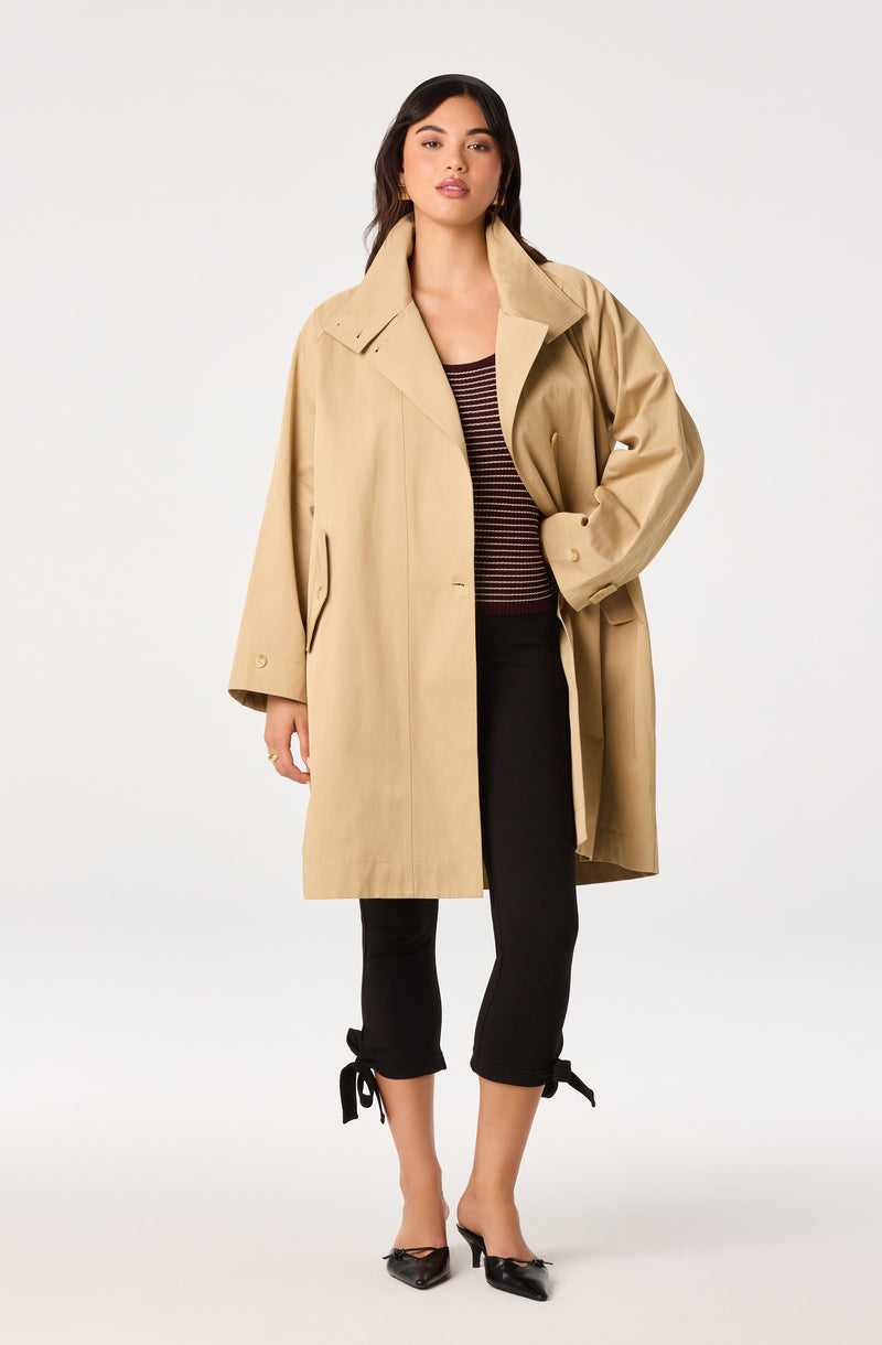 Woman wearing a beige trench coat, black cropped pants with bow ties, striped top, and black pointed shoes.
