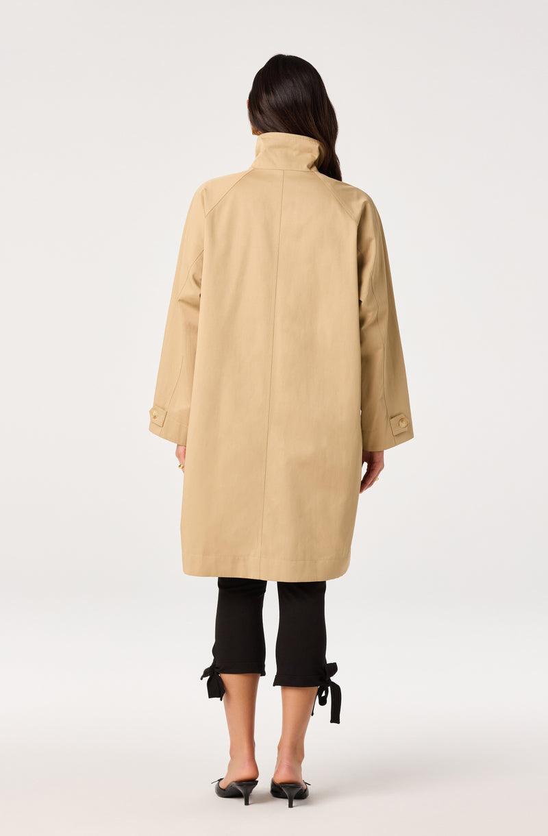 Beige knee-length coat with a high collar and buttoned cuffs, worn over black pants with tied hems.