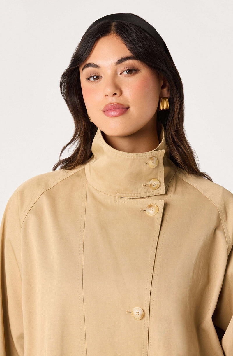 Woman in a beige high-collar coat with large buttons and gold earrings.