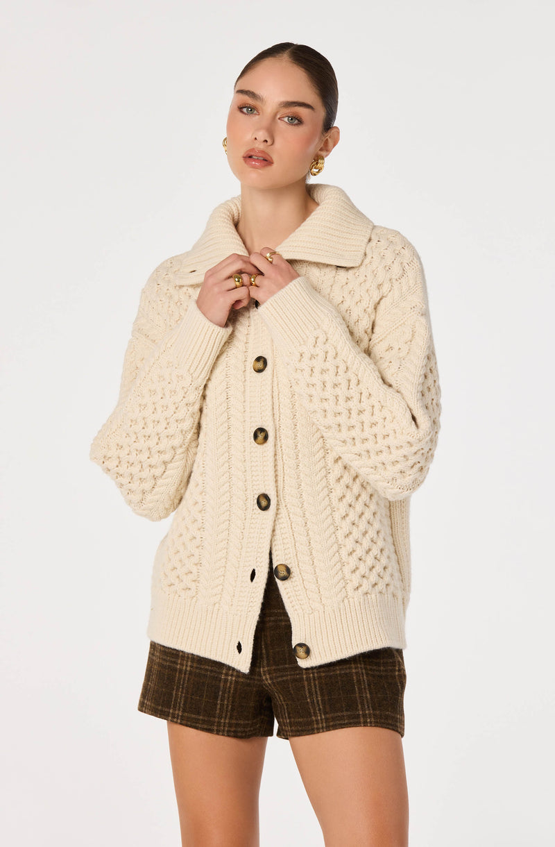 Woman wearing a cream cable-knit cardigan with large buttons and plaid shorts.