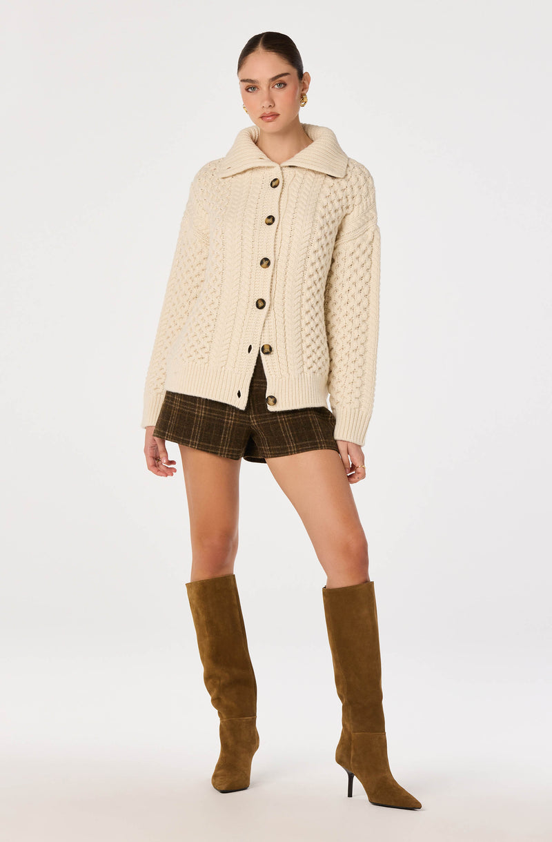 Cream chunky knit cardigan with large brown buttons, paired with dark plaid shorts and tall brown suede boots.