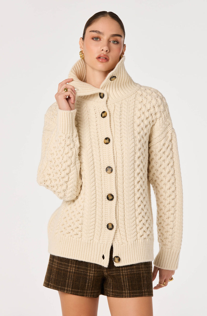 Woman wearing a cream chunky knit cardigan with large buttons and a high ribbed collar.