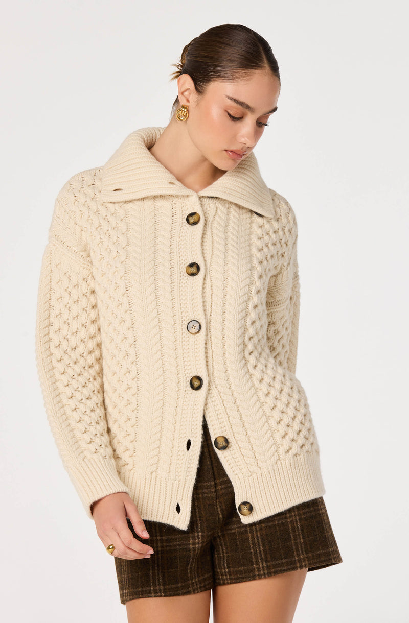 Woman wearing a cream cable-knit cardigan with large buttons and plaid brown shorts.