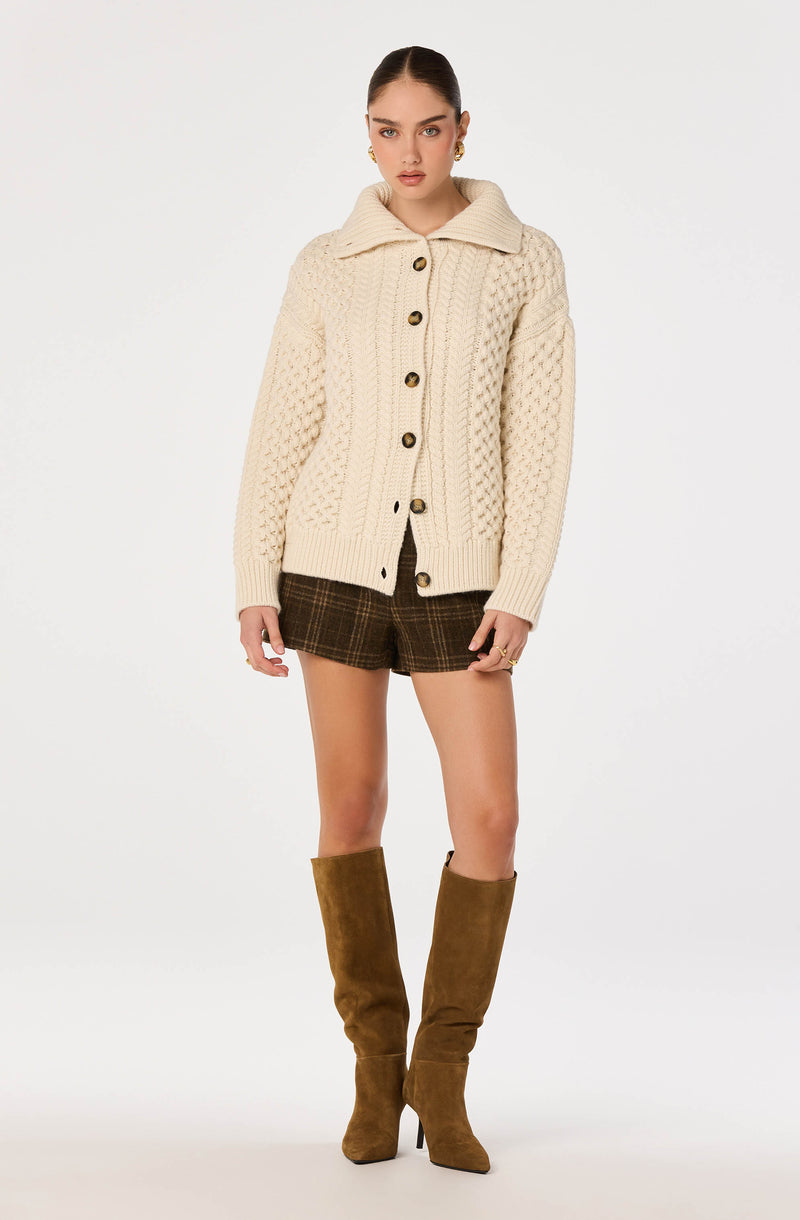 Woman wearing a chunky cream cable-knit cardigan, plaid shorts, and tall brown suede boots.