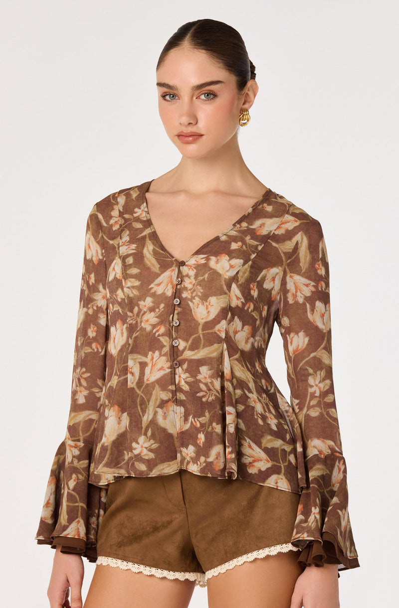 Woman wearing a brown floral print blouse with long ruffled sleeves and brown shorts.