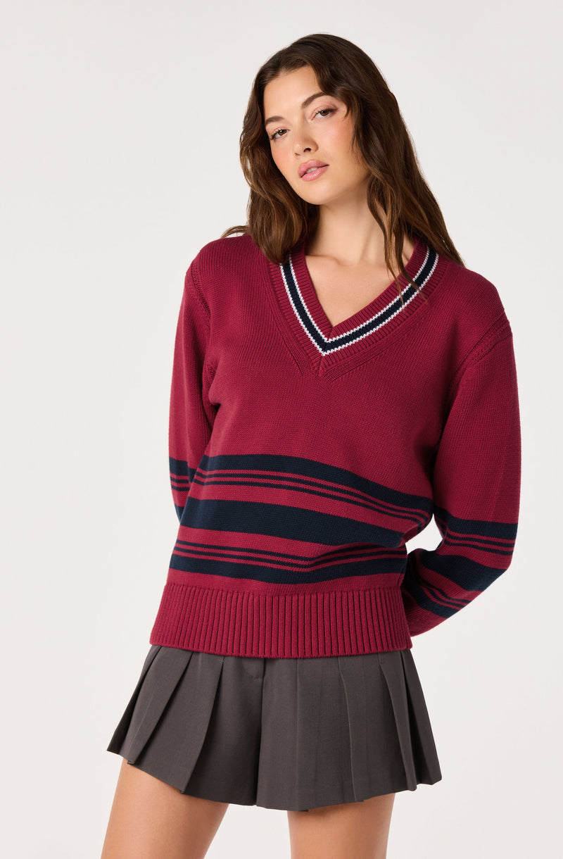 Girl wearing a red knit sweater with navy and white stripes and a pleated gray skirt.