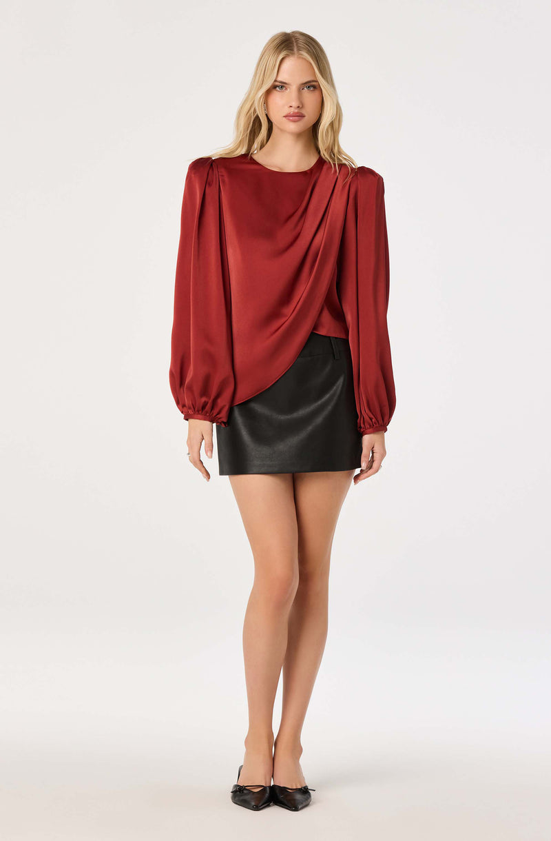 Woman in a red satin blouse with long, puffed sleeves and a draped front, paired with a black leather mini skirt and black pointed heels.