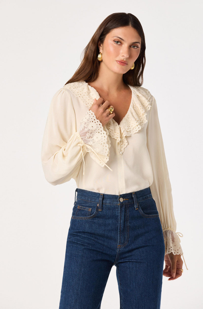Woman wearing a cream lace-embellished blouse and dark blue jeans.