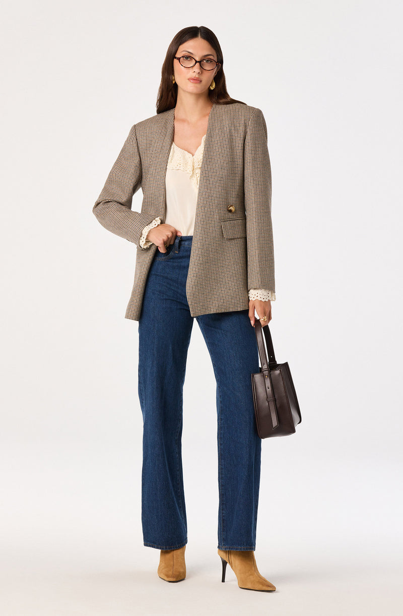 Woman wearing a plaid blazer, cream blouse with lace details, high-waisted blue jeans, beige high-heeled boots, and glasses, holding a black handbag.