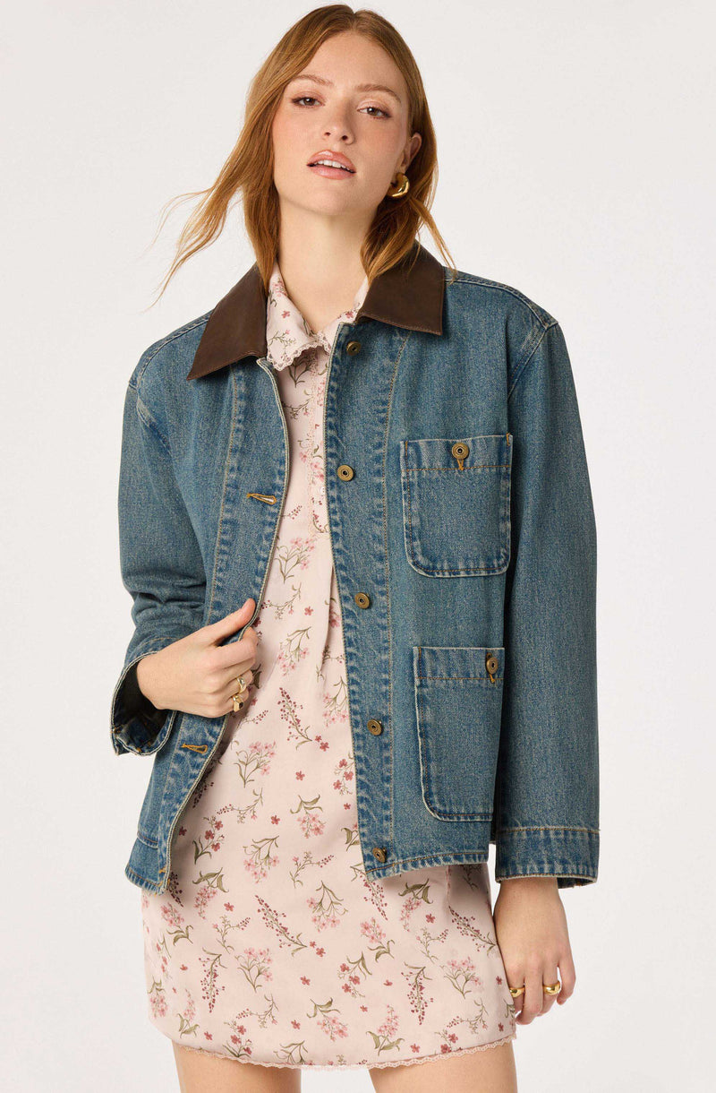 Woman wearing a denim jacket with a dark brown collar over a floral dress with pink and green pattern.