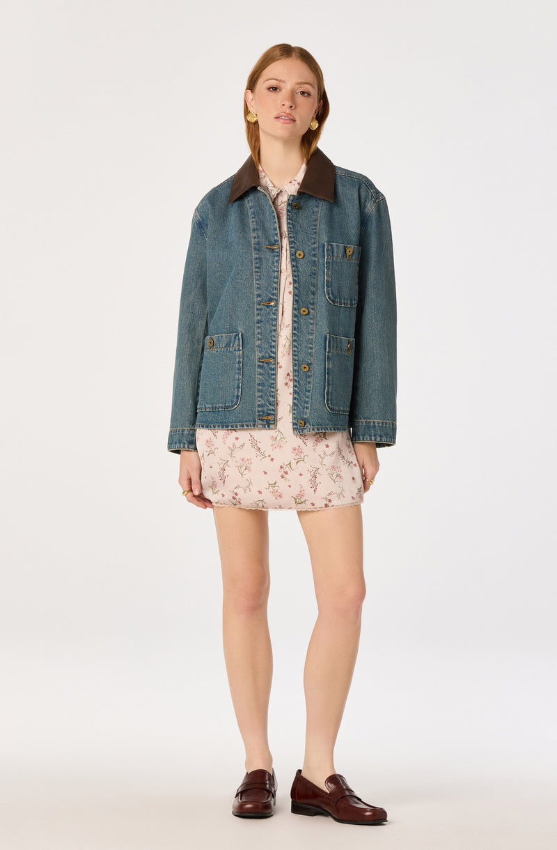 Woman wearing a denim jacket with a dark brown collar over a floral dress.