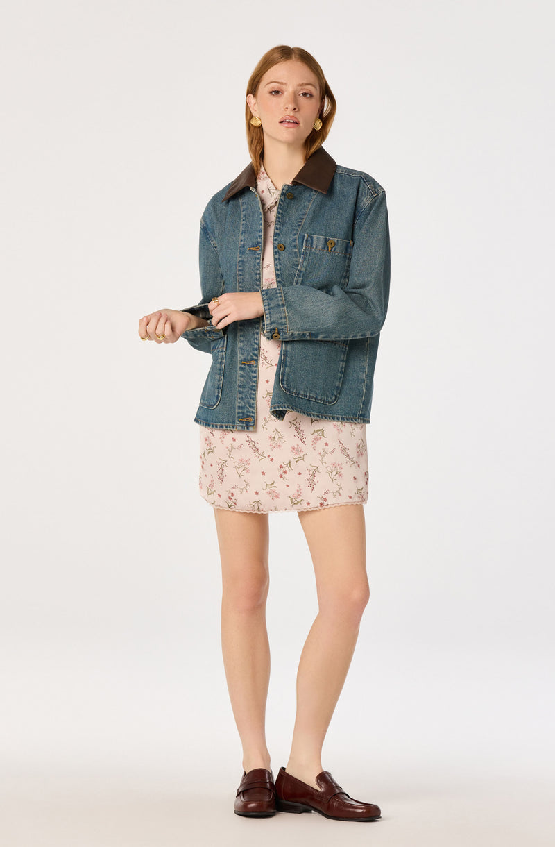 Woman wearing a blue denim jacket with brown collar, pink floral dress, and brown loafers.