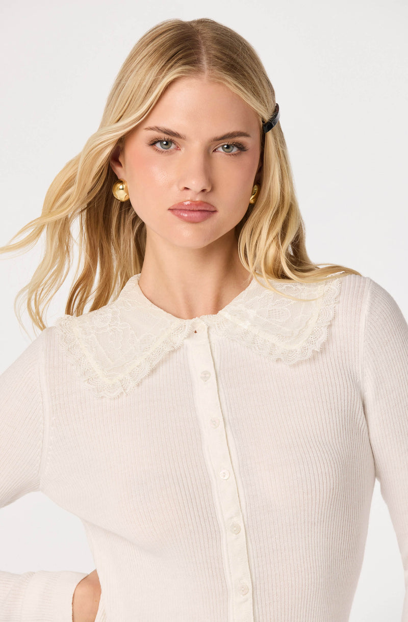 Woman wearing a cream-colored, ribbed knit top with a lace-trimmed collar, gold earrings, and a hair clip.