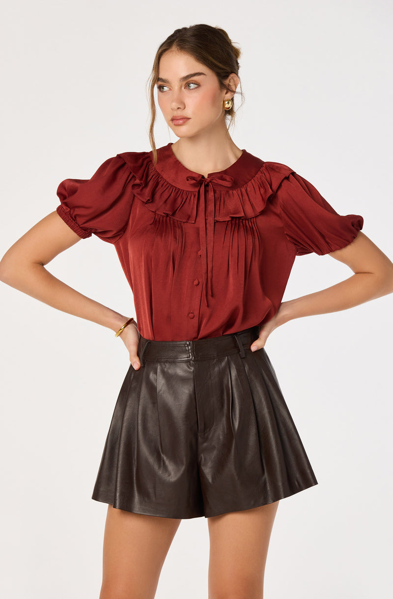 Woman wearing a silky red blouse with ruffle and pleats, paired with high-waisted black leather shorts.