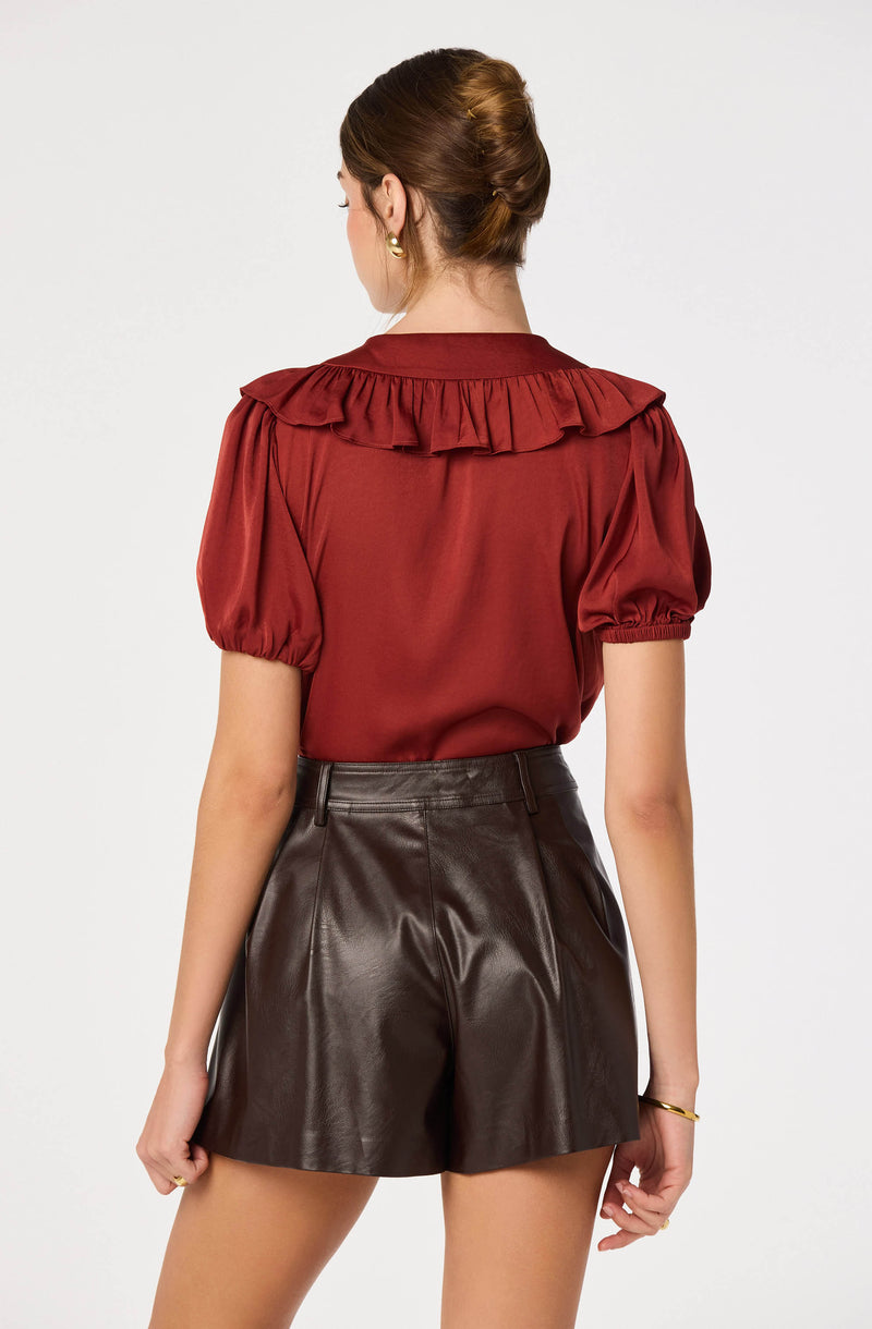 Woman wearing a rust-colored blouse with puffed short sleeves and ruffled collar, paired with dark leather shorts.