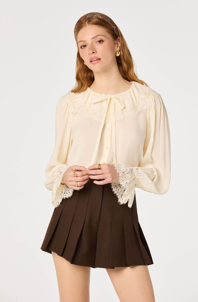 Woman wearing a cream lace-trim blouse with long sleeves and a pleated dark brown skirt.