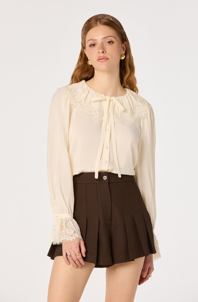 Woman wearing a cream blouse with lace details and dark pleated shorts.