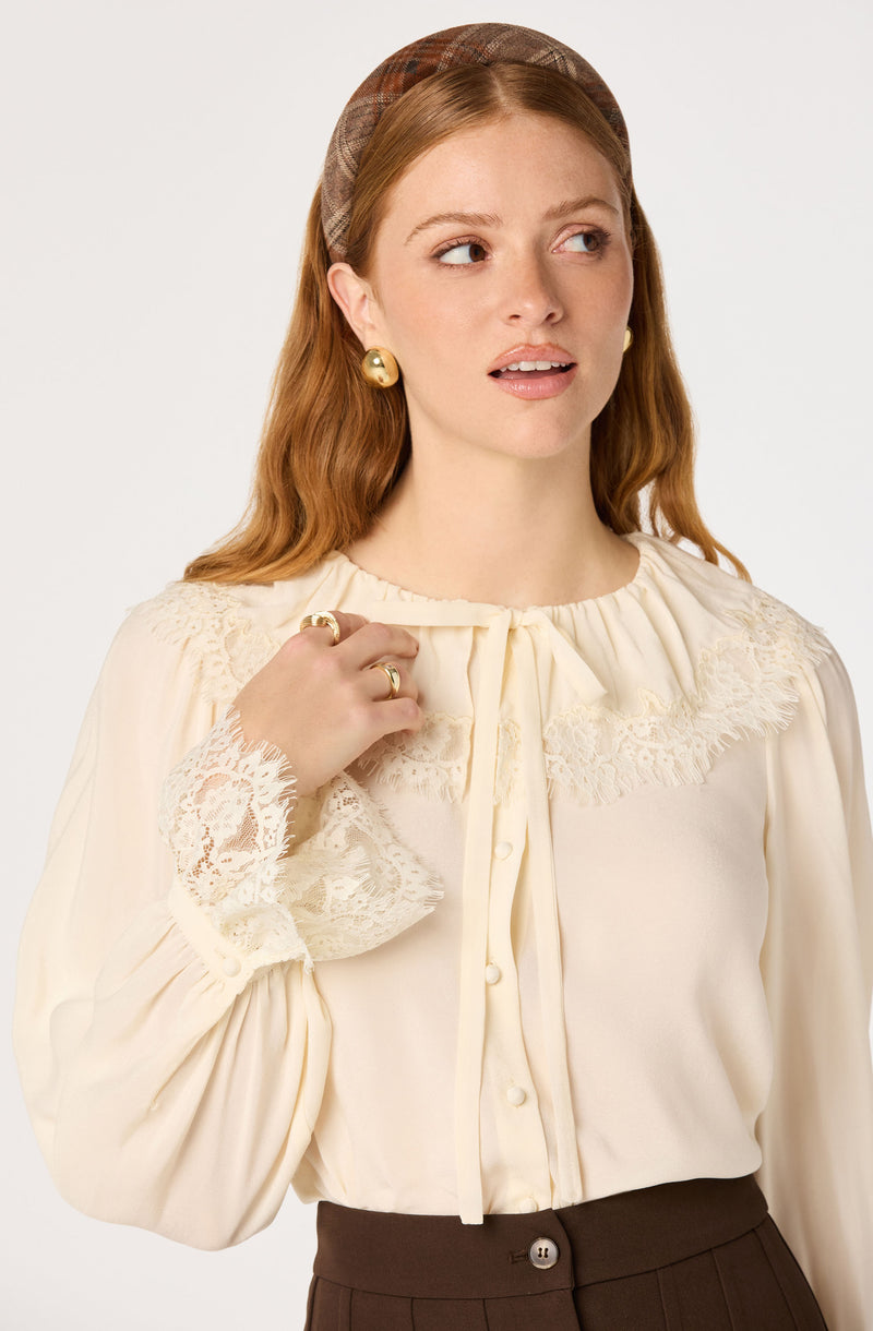 Woman wearing a cream blouse with lace details and gold accessories, paired with high-waisted brown pants.