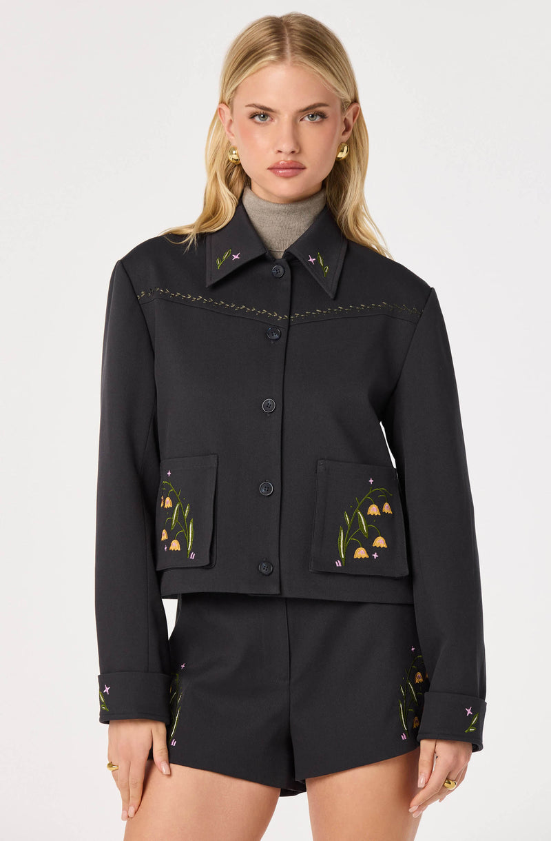 Black jacket with floral embroidered pockets, collar, and cuffs, worn over a gray turtleneck.