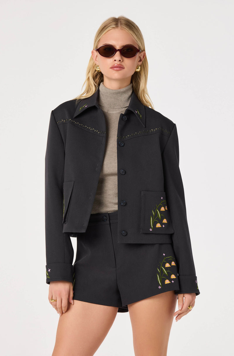 Woman in black jacket and shorts with floral embroidery, wearing sunglasses and gold jewelry.