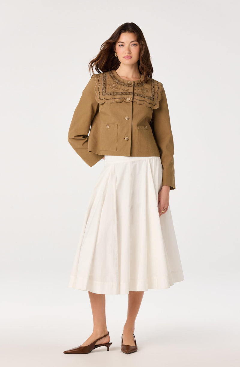 Woman wearing a tan jacket with embroidered details and a white, pleated midi skirt.