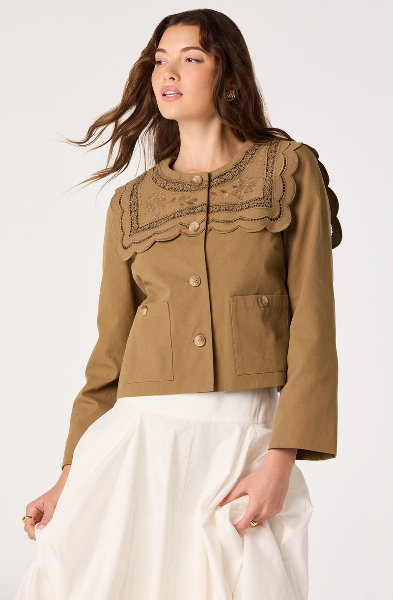 Woman wearing a tan jacket with embroidered lace collar and large front pockets.