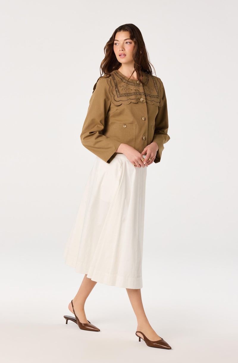 Model wearing a khaki jacket with embroidered details and cream pleated skirt, brown heeled shoes.