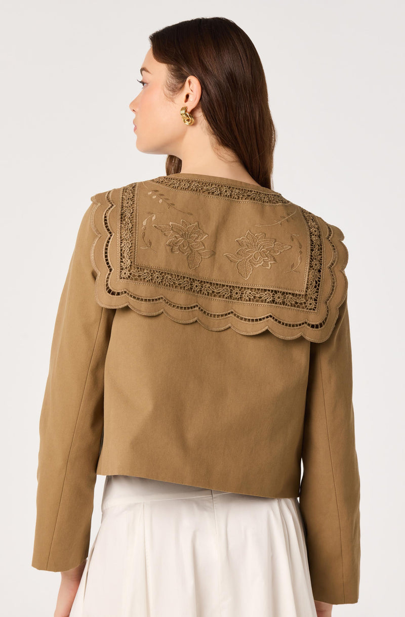 Woman in a tan jacket with embroidered floral design and scalloped edges on the shoulders.