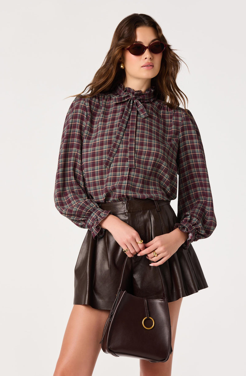 Woman in a plaid blouse with a bow neck, wearing dark sunglasses, and holding a black handbag.