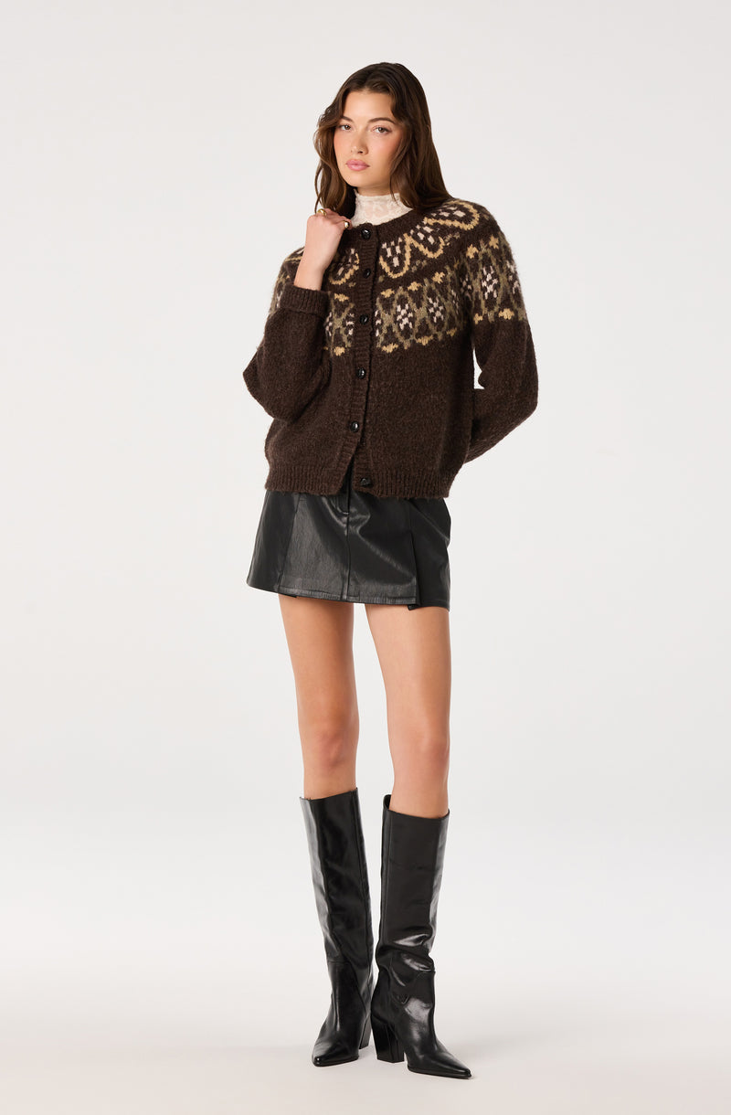 Model wearing a patterned brown cardigan, black leather mini skirt, and tall black boots.