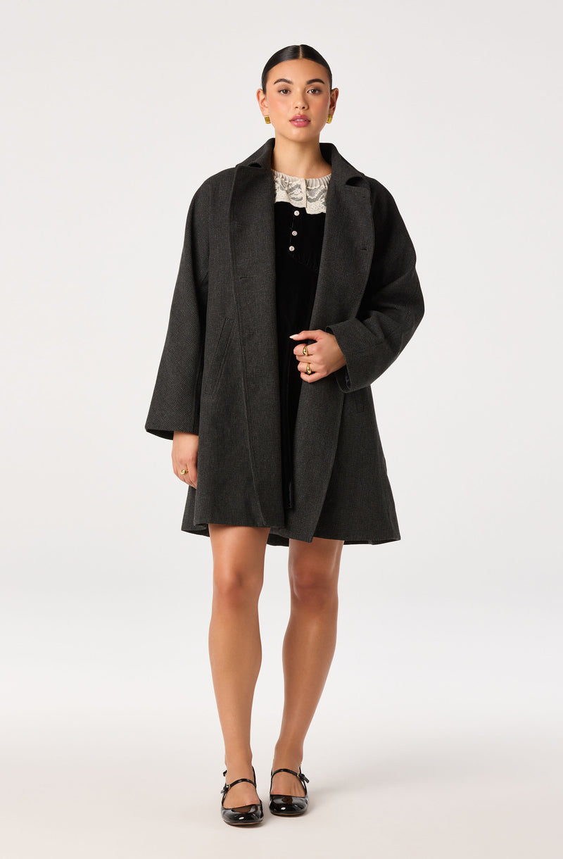 Woman wearing a dark gray coat over a black dress with a lace collar, black heels with ankles straps.