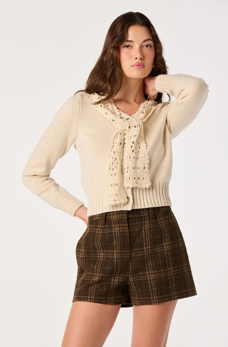 Woman wearing a cream knit sweater with a lace scarf detail and brown plaid shorts.