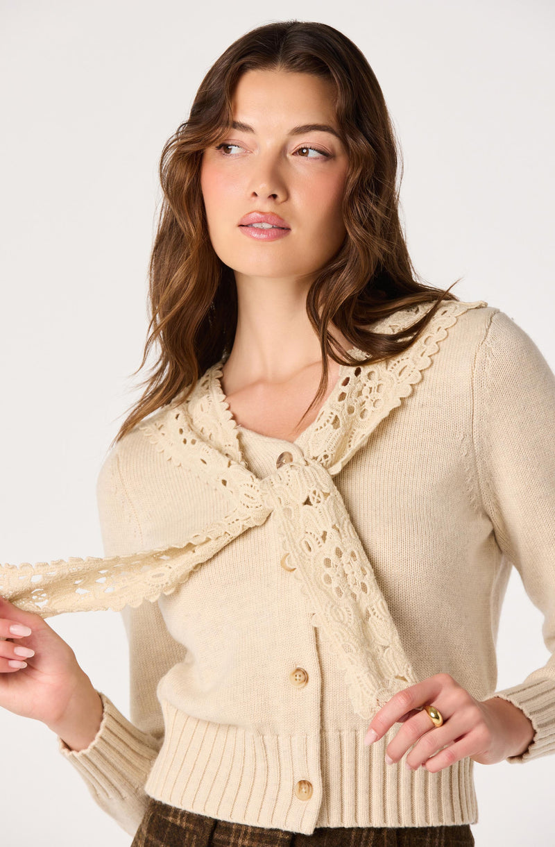 Woman wearing a beige knit cardigan with a scalloped lace collar and matching lace tie.