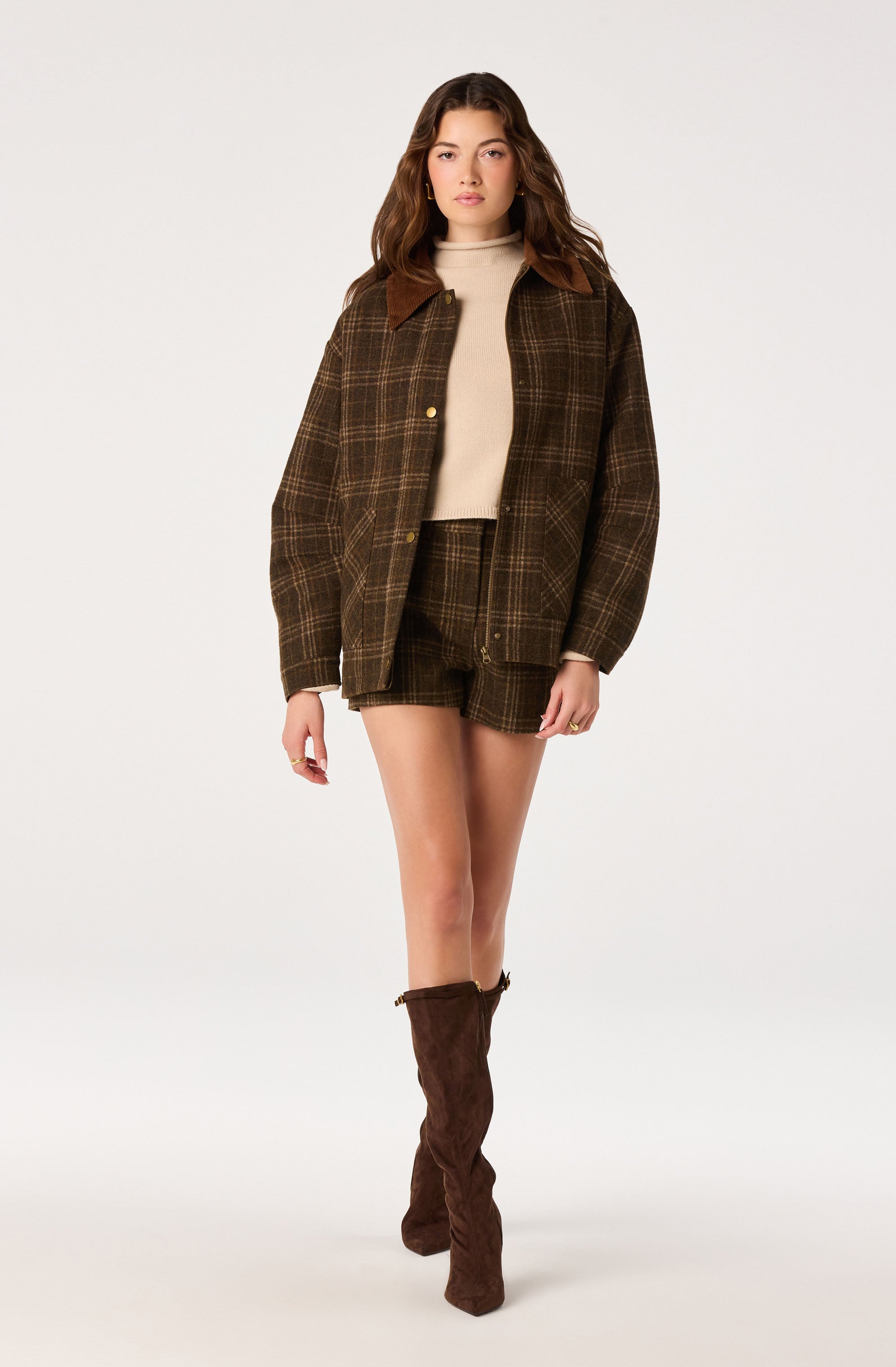 Woman wearing a brown plaid jacket with brown suede collar, matching plaid shorts, beige turtleneck sweater, and brown knee-high boots.