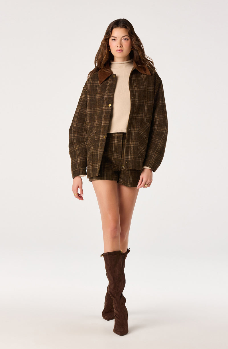 Woman wearing a brown plaid jacket with brown suede collar, matching plaid shorts, beige turtleneck sweater, and brown knee-high boots.