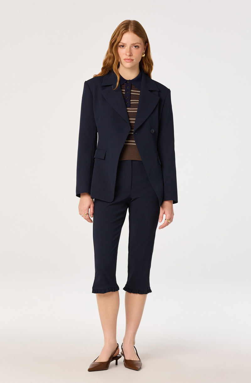 Woman in a navy blazer, dark pants with ruffled hem, striped shirt, and brown heels.