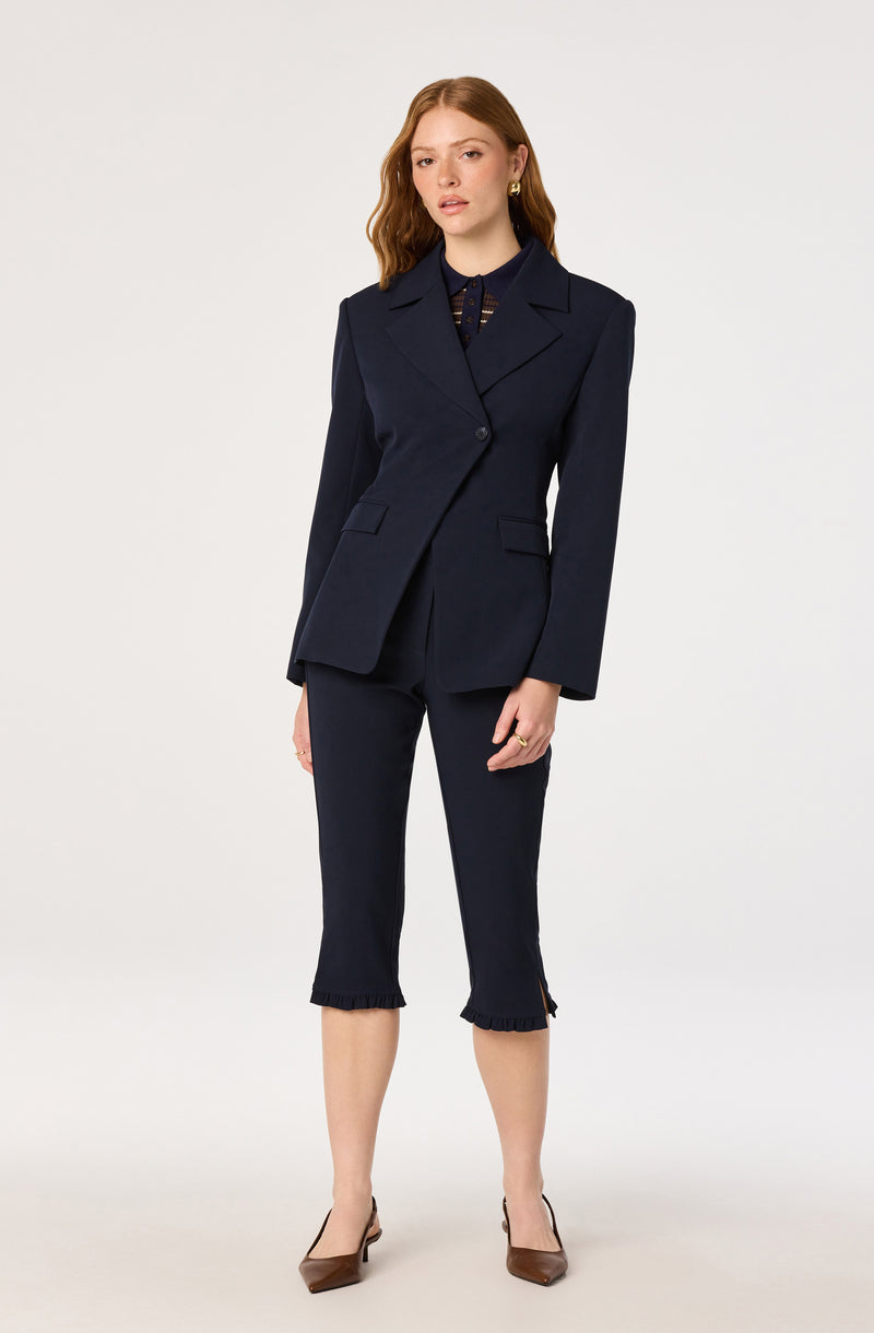 Woman wearing a navy tailored blazer and cropped pants with ruffled hems, brown heels.