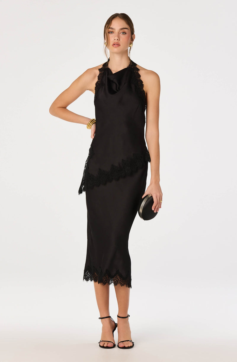 Woman in black satin dress with lace trim, sleeveless, midi-length, with a draped neckline, holding a small black clutch.