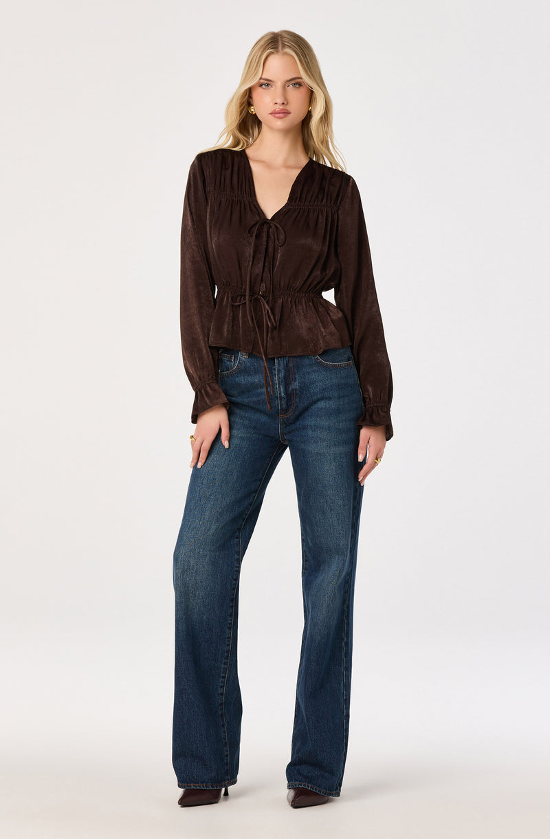 Woman in a brown, long-sleeve blouse with a tie-front detail, paired with blue jeans.