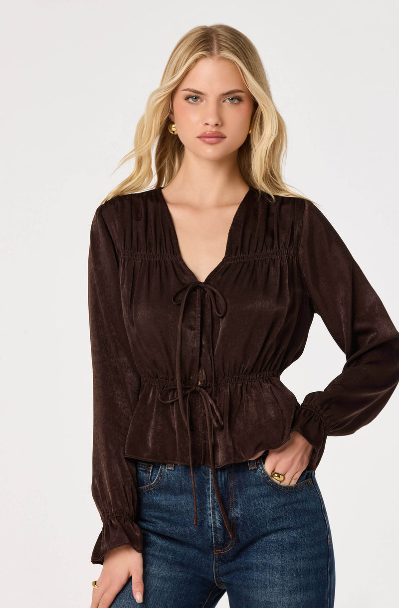 Woman wearing a dark brown, long-sleeve blouse with ruffled shoulders and tie-front details.