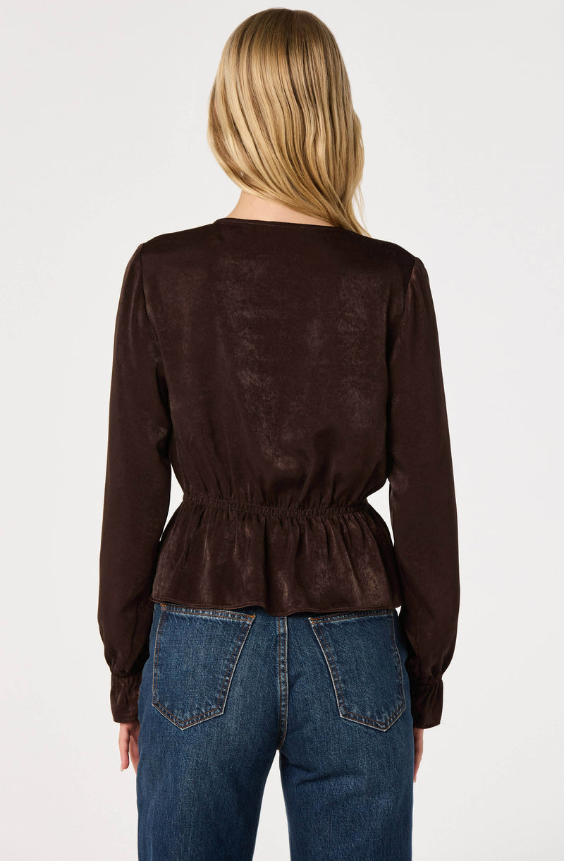 Woman wearing a dark brown, long-sleeve, cinched waist satin blouse with a peplum hem.
