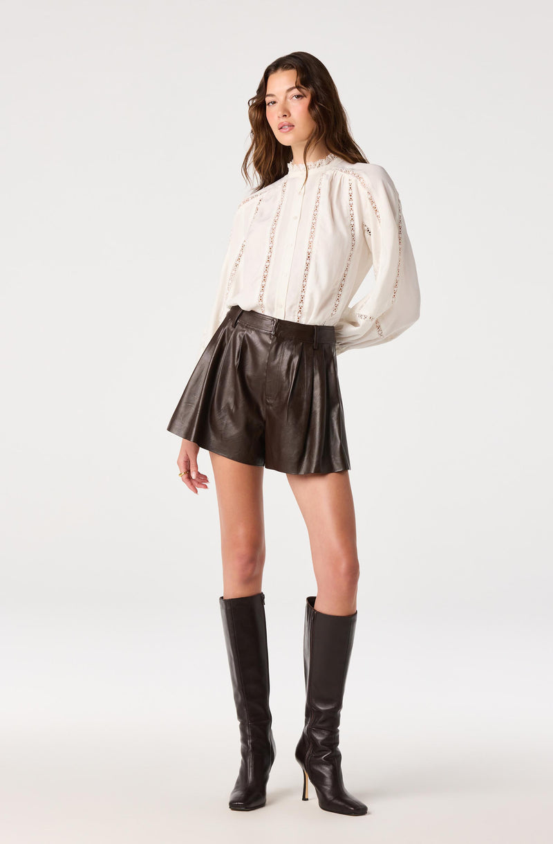 Woman wearing a white long-sleeve blouse with lace details and high collar, paired with black leather shorts and knee-high black leather boots.
