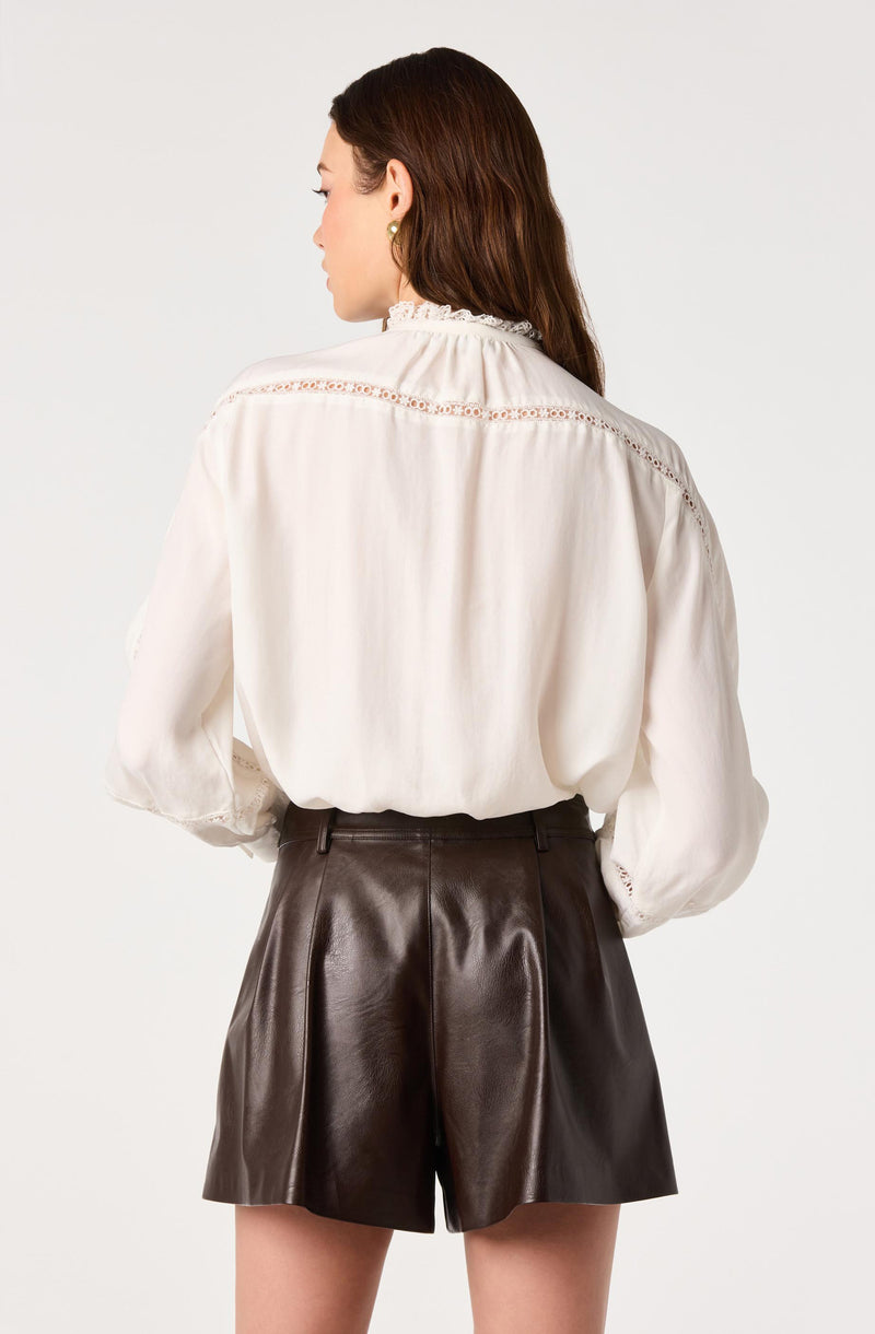Woman wearing a white blouse with lace details and high-waisted brown leather shorts.