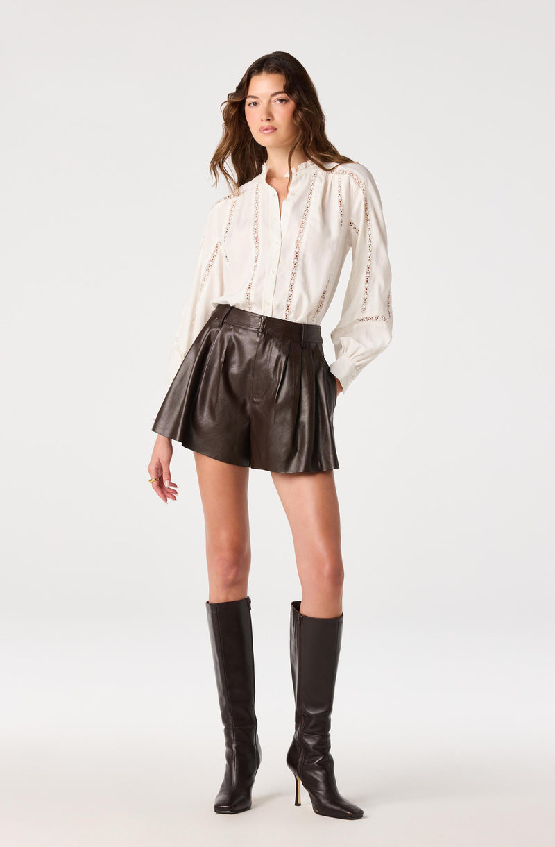 Woman wearing a white embroidered blouse, dark brown leather shorts, and tall black leather boots.