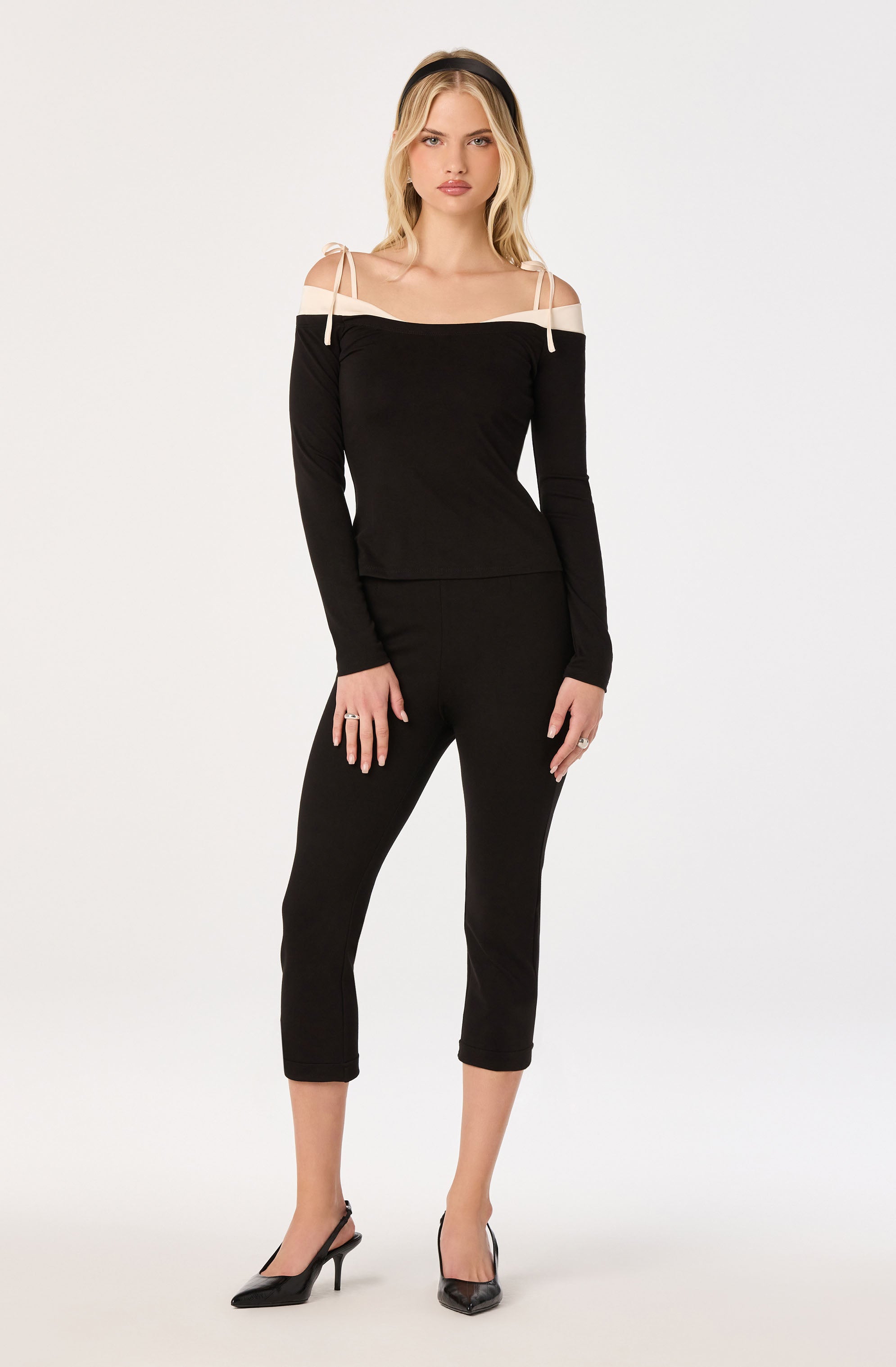 Woman wearing a black long-sleeve top with off-shoulder beige cutouts and tie straps, paired with black ankle-length pants.
